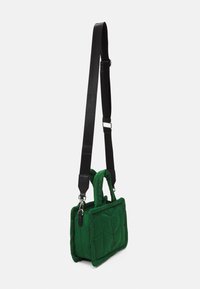 Sac à main carré matelassé vert avec des poignées courtes et une longue bandoulière noire réglable suspendu sur un fond blanc.