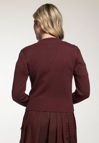 Burgunderfarbener Strickcardigan mit gerippter Textur und gewelltem Ausschnitt; verfügt über ein dekoratives Herzmuster auf dem Rücken.