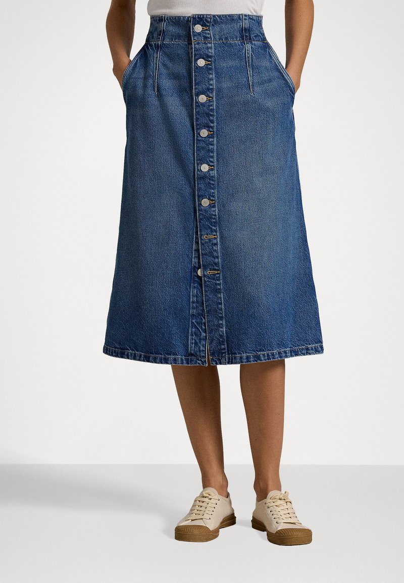 Denim midi-hame, jossa on tiukka vyötärö, etupainike, sivutaskut ja hieman repaleinen helma keskisinisessä värissä. Yhdistettynä beigeksi lenkkareihin.