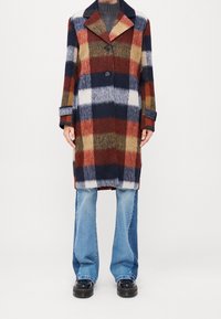 Cappotto in plaid di lana nei colori blu navy, rosso, beige e bianco, con un ampio colletto, tre bottoni e maniche a contrasto; abbinato a jeans svasati di un leggero blu.