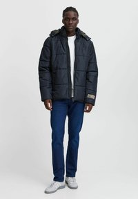 Svart puffjakke med avtagbar hette, glidelås-lukking og quiltet design. Båret over en hvit t-skjorte og kombinert med blå jeans og grå joggesko.