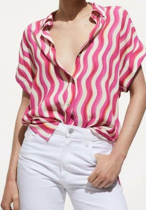 Osoba ubrana w krótki rękaw, różowo-białą koszulę w fale z guzikami, włożoną do białych spodni z wysokim stanem, ręka spoczywa blisko kieszeni.