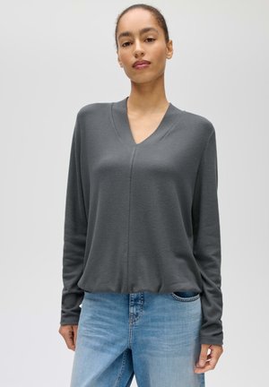 Femme portant un haut à manches longues gris foncé à col en V et un jean bleu clair, debout devant un fond gris clair uni.