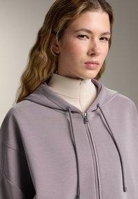 Felpa zip-up lilla con lacci regolabili, texture morbida e cappuccio grande, indossata sopra un top beige a collo alto attillato.