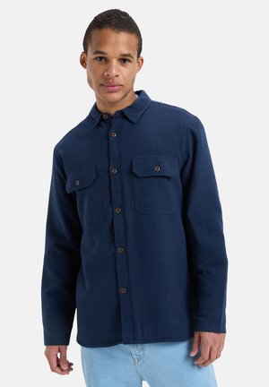 Chemise bleu marine à manches longues, en tissu texturé. Présente deux poches poitrine et des boutons bruns. Coupe avec un col classique.