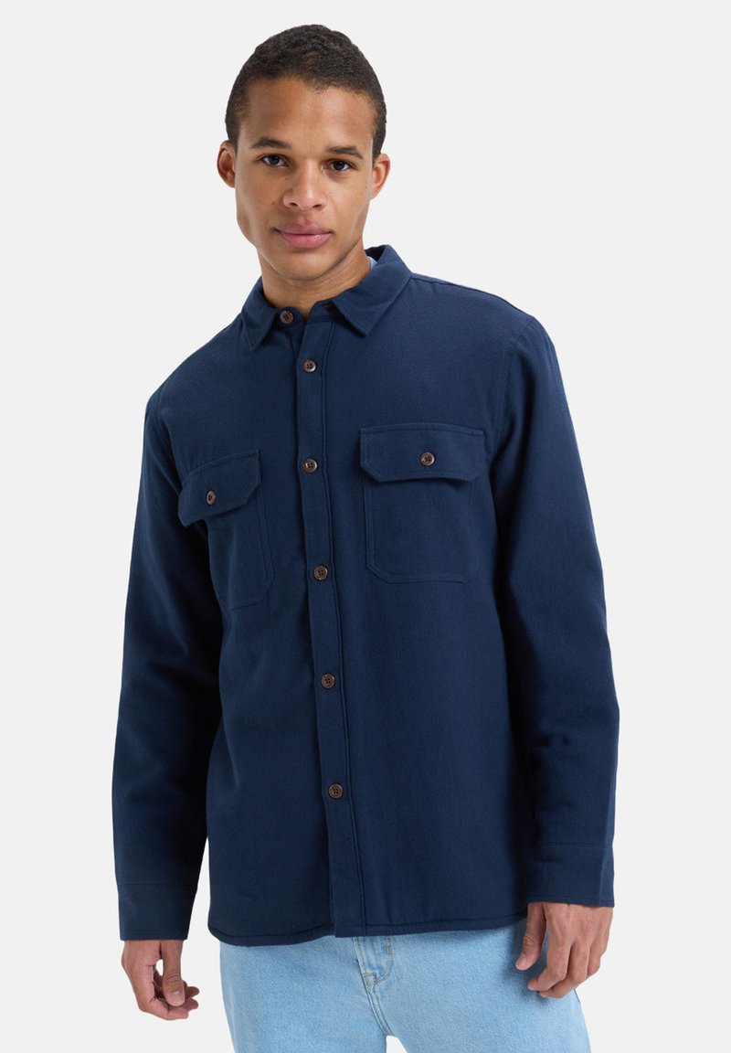 Marineblauwe button-up shirt gemaakt van textuurstof. Heeft twee borstzakken en bruine knopen. Geknipt met een klassieke kraag en lange mouwen.