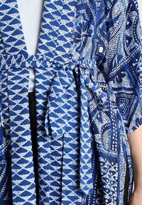 Niebiesko-biała szata z wzorem geometrycznym i kwiatowym, przewiązana w talii nad białą bluzką i czarnymi spodniami.