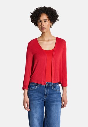 Rote, leichte Strickjacke mit Rundhalsausschnitt und offenen Front, kombiniert mit einem roten Trägershirt darunter. Blaue Denim-Jeans runden das Outfit ab.