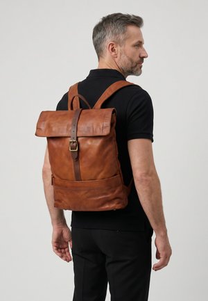Man draagt een zwarte polo en broek met een bruine leren rugzak met voorste riem en gesp, zijwaarts kijkend tegen een lichtgrijze achtergrond.