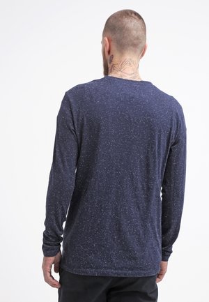 Mann mit Hals-Tattoos, der ein marineblaues, gesprenkeltes Langarmshirt trägt und mit dem Rücken zur Kamera vor einem schlichten weißen Hintergrund steht.