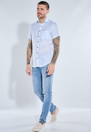 Mann mit tätowierten Armen, der ein weißes kurzärmeliges Hemd mit Knöpfen, hellblaue Jeans und beige Slipper trägt, steht vor weißem Hintergrund.