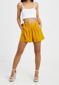 Żółte szorty o wysokim stanie z paskiem do wiązania, wykonane z lekkiego materiału. Połączone z białym topem na ramiączkach i sandałami na platformie.