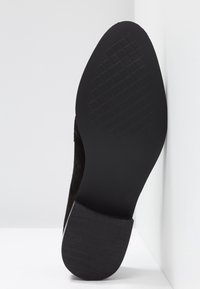 Semelle de chaussure noire avec une zone texturée à l'avant et un talon lisse légèrement surélevé, positionnée sur un fond blanc en angle.