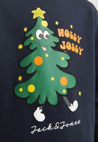 Zöld grafikai pulóver, amelyen egy rajzfilmszerű karácsonyfa, sárga csillag, narancssárga körök és a "HOLLY JOLLY" felirat látható tengerészkék háttéren.