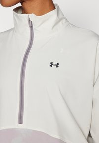Svetlo siva zip-up jakna z visokim ovratnikom, opremljena z vijoličnim zadrgo in črnim logotipom. Gladka tekstura, zasnovana za udobje in mobilnost.