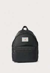 Mochila de tecido preto com forma arredondada, apresentando um bolso frontal com fecho de correr e um emblema com o logótipo. Alças ajustáveis e pega superior para transporte.