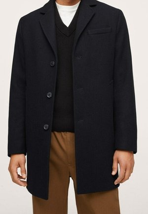 Mann trägt einen schwarzen, geknöpften Mantel über einem schwarzen V-Ausschnitt-Pullover und weißem Hemd, kombiniert mit braunen Hosen, dargestellt von den Schultern bis zur Mitte des Oberschenkels.