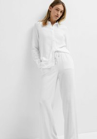 Witte, lichte button-up shirt met lange mouwen, gecombineerd met bijpassende joggingsbroek met trekkoord. Beide hebben een gladde, gestructureerde stof.