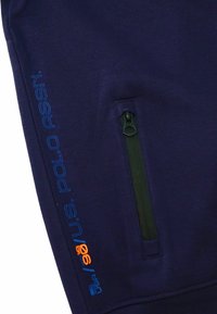 Námořnicky modrá tkanina s černou zipovou kapsou a modro-oranžovou značkou "U.S. POLO ASSN." a textem "USA 96" po straně.