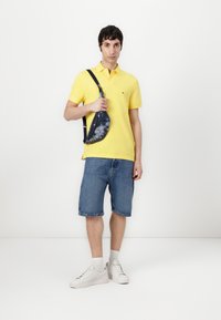 Cămașă polo galbenă, șorturi din denim bleu, adidași albi, șosete gri. Modelul ține o geantă crossbody cu un model tie-dye albastru, cu o curea neagră.