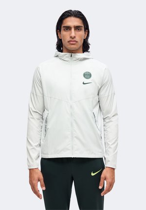 Homme portant une veste à capuche gris clair avec les logos Paris Saint-Germain et Nike, et un pantalon noir avec le logo jaune Nike.
