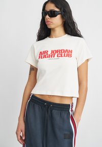 Δερματινή κρεμ t-shirt με τη γραφή "AIR JORDAN FLIGHT CLUB" σε κόκκινη εκτύπωση, συνδυασμένη με σκούρα φόρμα που διαθέτει λευκές και κόκκινες ρίγες κατά μήκος των πλαισίων.