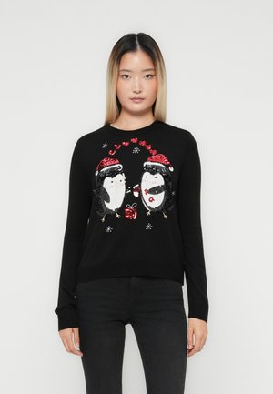 Mulher vestindo um suéter preto com dois pinguins brilhantes em chapéus de Natal trocando um presente, cercados por bengalas de doces e flocos de neve.