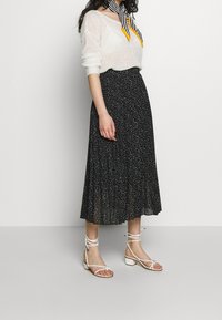 Schwarzer plissierter Midirock mit weißen Polka-Dots, kombiniert mit einem leichten weißen Pullover und weißen Riemchensandalen, der einen lässigen Look präsentiert.