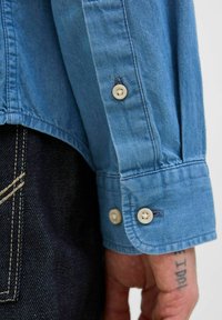 Ljusblå denimskjorta med ärm som har två gräddvita knappar, synlig sömnad och subtil textur. Mörka jeans delvis synliga nedanför.