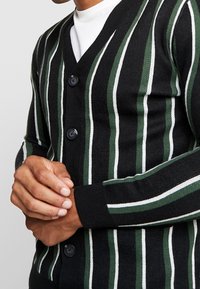 Cárdigan negro con rayas verticales en verde y blanco, con cuello en V, cinco botones y puños acanalados para un acabado texturizado.