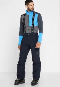 Giacca con zip blu e grigia con colletto alto, abbinata a pantaloni da sci blu scuro con bretelle regolabili e tessuto strutturato.