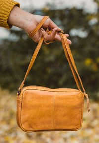 Hand som bär en senapsgul tröja håller en liten beige lädercrossbody-väska i sina remmar framför en suddig utomhusbakgrund.