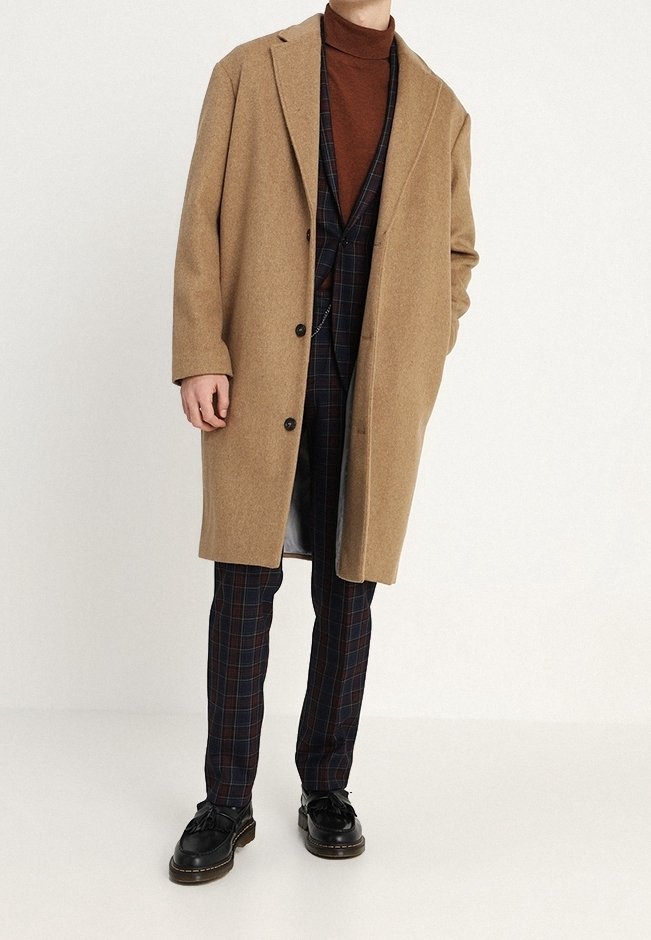 Uomo con un cappotto color cammello, dolcevita marrone, completo a quadri blu navy e scarpe di pelle nere che posa su uno sfondo neutro.