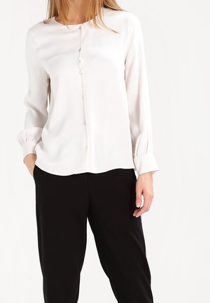 Kvinde iført en langærmet hvid knapbluse med manchetter, kombineret med sorte løstsiddende bukser.