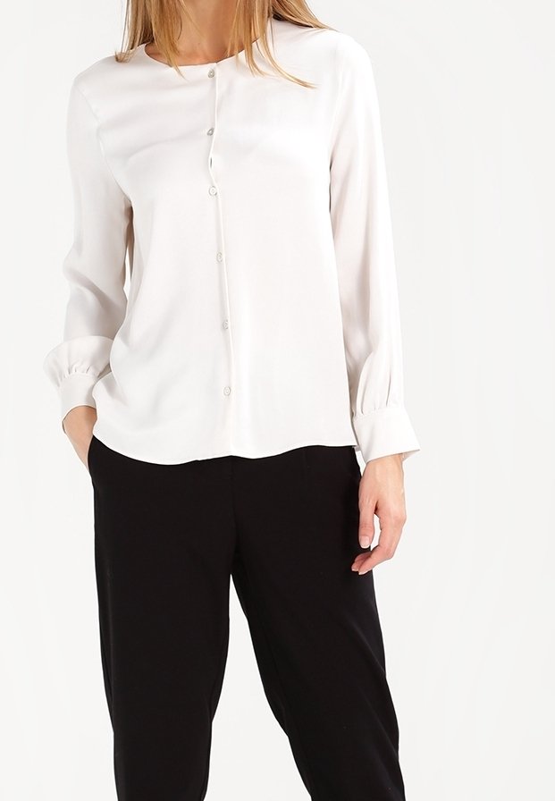 Femme portant un chemisier blanc à manches longues boutonné avec des manches retroussées, associé à un pantalon noir ample.