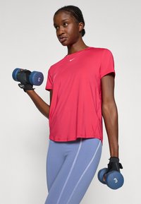 Camiseta deportiva roja de manga corta, con logo en el pecho; mallas moradas con rayas laterales, sosteniendo mancuernas azules con el número 3 en ellas.