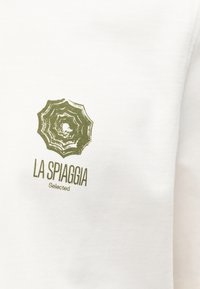 Λευκό ύφασμα με πράσινο γραφικό μιας παραλίας ομπρέλας και το κείμενο "LA SPIAGGIA Selected" τυπωμένο από κάτω.