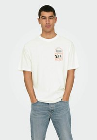 Camiseta blanca de algodón con un gráfico frontal de una hamburguesa y personajes de dibujos animados. Mangas cortas y un ajuste relajado. Combinada con unos vaqueros de color azul claro.