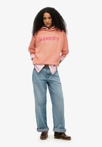 Roze hoodie met 'Superdry' logo, gecombineerd met een lichtroze shirt, ontspannen blauwe jeans en bruine schoenen. Krullend haar dat het gezicht frames.