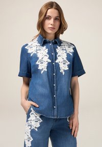 Camicia di jeans a maniche corte, con ricami floreali bianchi sulle spalle e bottoni sul davanti. Vestibilità tailored con tasca laterale.