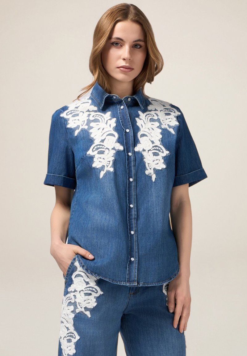 Camicia di jeans a maniche corte, con ricami floreali bianchi sulle spalle e bottoni sul davanti. Vestibilità tailored con tasca laterale.