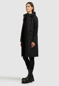 Kobieta stojąca w długim czarnym kapturowym płaszczu, czarnych leginsach i masywnych czarnych butach na tle jasnego, jednolitego tła.