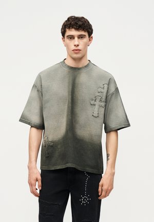 Ung mand med kort krøllet hår iført en oversized falmet olivengrøn t-shirt med slidte korspatches og sorte revnede jeans med nitte-detaljer.