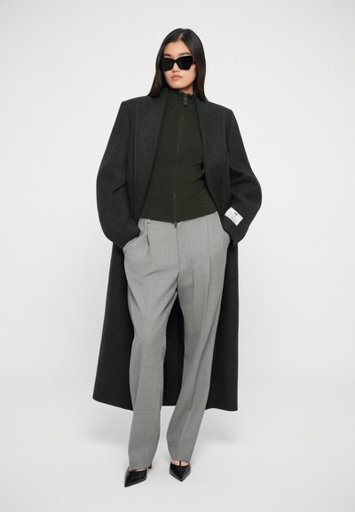 Calças cinzentas ajustadas com um suéter verde escuro com fecho e um casaco de lã preto oversized, combinados com saltos pretos de bico e óculos de sol.
