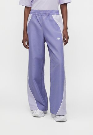 Person iført løse lavendel- og hvide træningsbukser med elastisk talje og hvide sneakers, stående mod en ensfarvet baggrund.