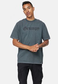 Camiseta de algodón gris con un estampado negro de "Grumpy" en una fuente audaz, de manga corta y ajuste holgado. Detalle de costura notable en el dobladillo.