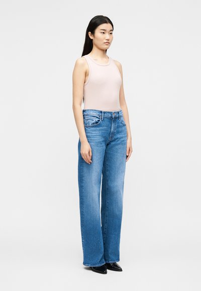 Νεαρή γυναίκα που φοράει ανοιχτό ροζ αμάνικο τοπ, ψηλόμεσα μπλε φαρδιά τζιν και μαύρα μυτερά παπούτσια, στέκεται μπροστά σε λευκό φόντο.