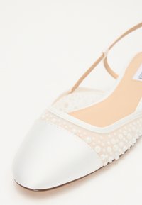 Zapato plano de satén blanco con punta afilada, parte superior transparente adornada con pequeñas perlas blancas y una delicada correa en el tobillo.