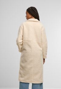 Helle beige, flauschige Wolljacke mit Kragen, langen Ärmeln und lockerer Passform. Nahtdetails entlang des Rückens und weiche Textur.