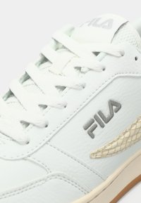 Hvit lær sneaker med grå Fila-logo, teksturerte detaljer og flate hvite lisser. Sålen har en beige gummibunn for grep.